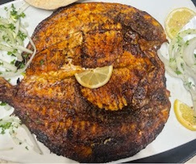 Whole grilled fish, garnished with lemon slices, chopped parsley, and sliced onions.